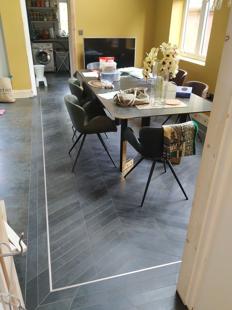 Ułożona pod kątem ciemnoszara podłoga z płytek, wykończona srebrną listwą przypodłogową, w tle stół z krzesłami w jadalni.