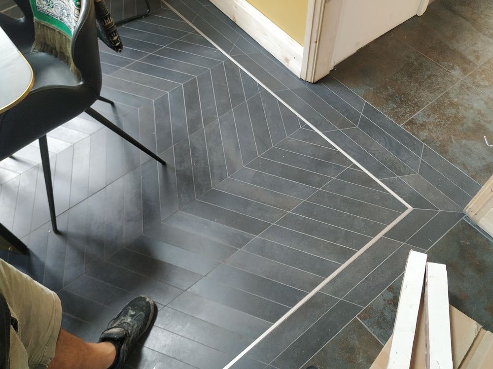 Ułożona w jodełkę, grafitowa podłoga z płytek ceramicznych, oddzielona listwą od starej podłogi, widoczne fragmenty krzesła i roboczych materiałów.