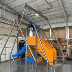 Konstrukcja stalowa placu zabaw z pomarańczową zjeżdżalnią i niebieskimi elementami, wykonana ze stali nierdzewnej, w hali produkcyjnej z widocznymi narzędziami na ścianie.