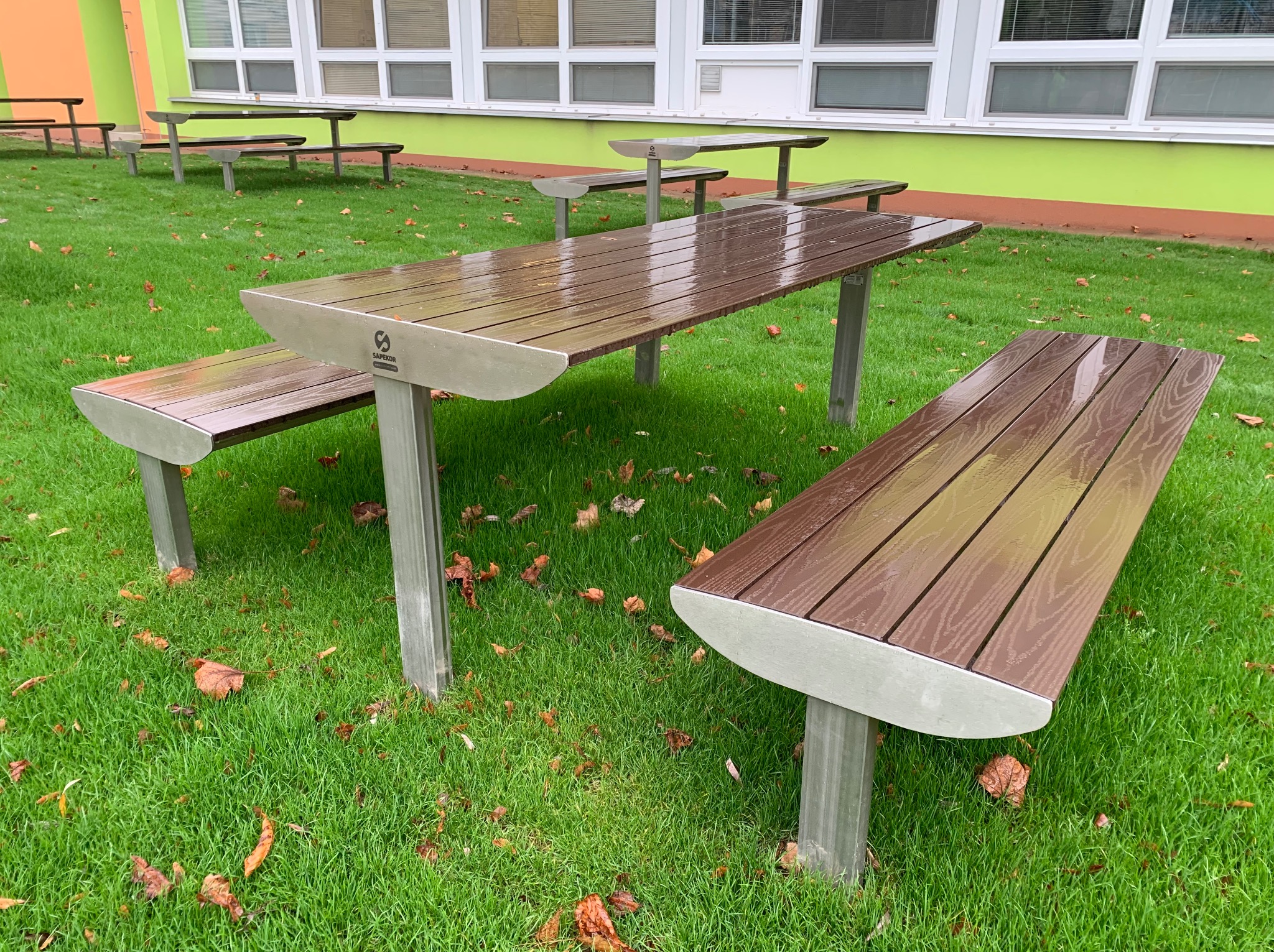 Zestaw mebli miejskich: stół i ławki z brązowego drewna kompozytowego na metalowych nogach, ustawione na trawniku z opadłymi liśćmi, widoczne z bliska na tle budynku z oknami.