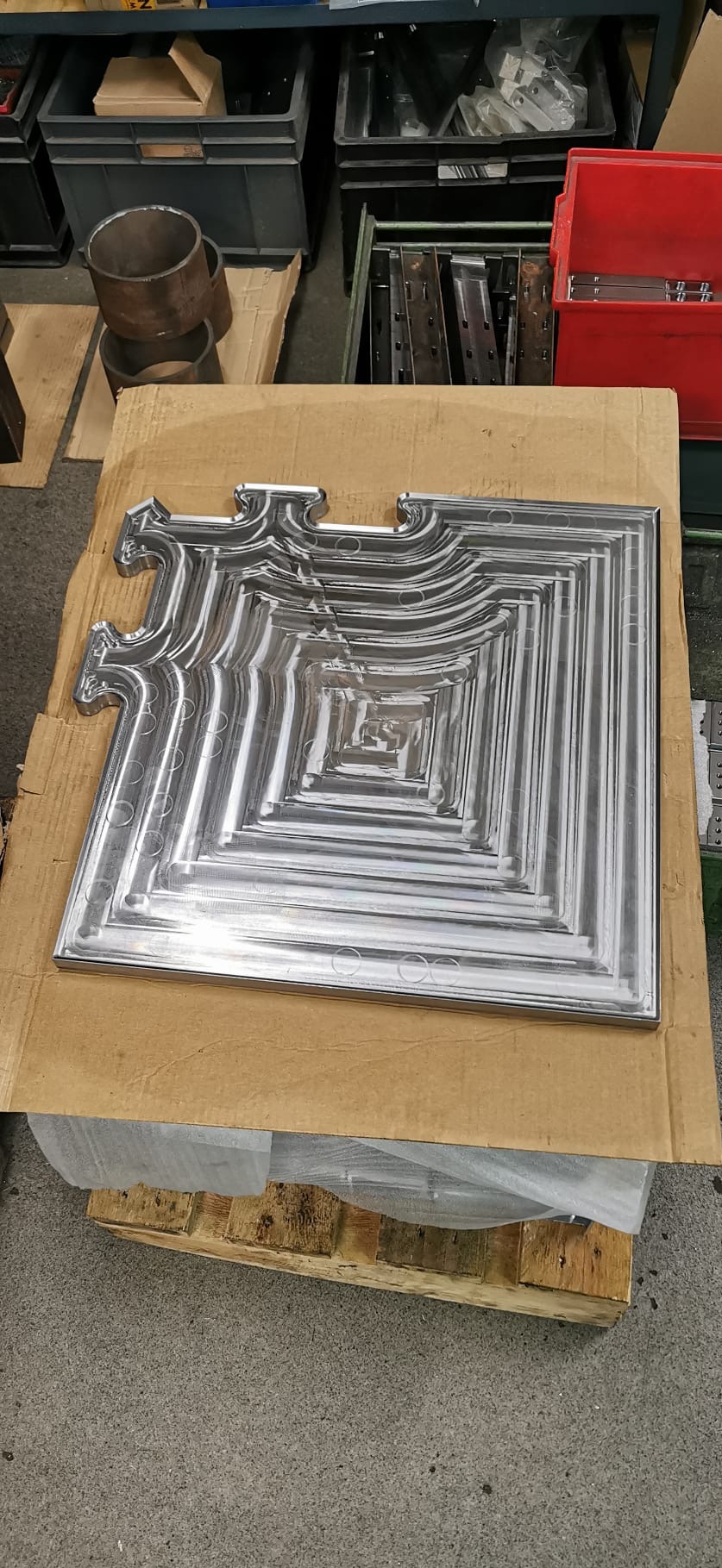 Błyszczący, metalowy element dekoracyjny z powtarzającym się motywem kwadratów, leżący na tekturowym podłożu w warsztacie.