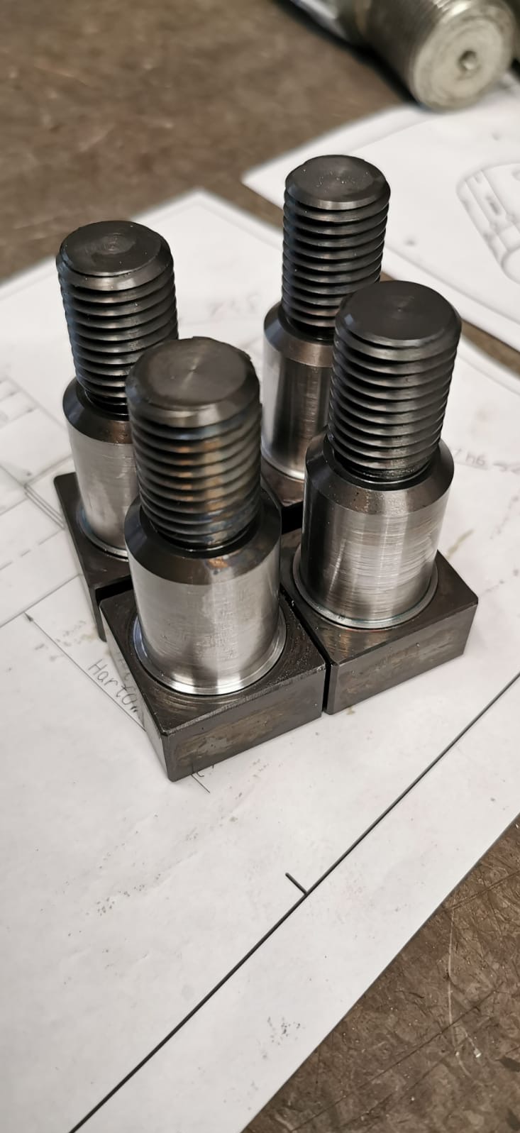 Cztery metalowe śruby z gwintem i kwadratową podstawą, ułożone na rysunku technicznym z widocznymi liniami i fragmentem napisu 'Harton', z bliska, w warsztatowym otoczeniu.