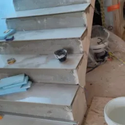 Schody w trakcie wykańczania z użyciem poziomicy laserowej, widoczne płytki, zaprawa, narzędzia i akcesoria montażowe.