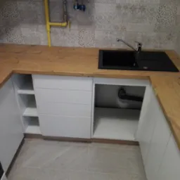 Nowoczesna kuchnia z drewnianym blatem, czarnym zlewem i białymi szafkami, widoczna żółta rura gazowa na tle wzorzystych płytek.