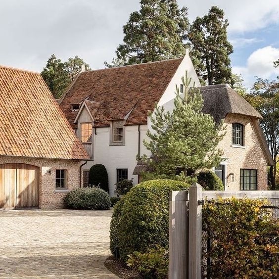 Urokliwy dom z dachem krytym strzechą i ceglanym dachem garażu, otoczony zielenią. Jasna elewacja, kamienny podjazd i drewniane ogrodzenie dodają rustykalnego charakteru.