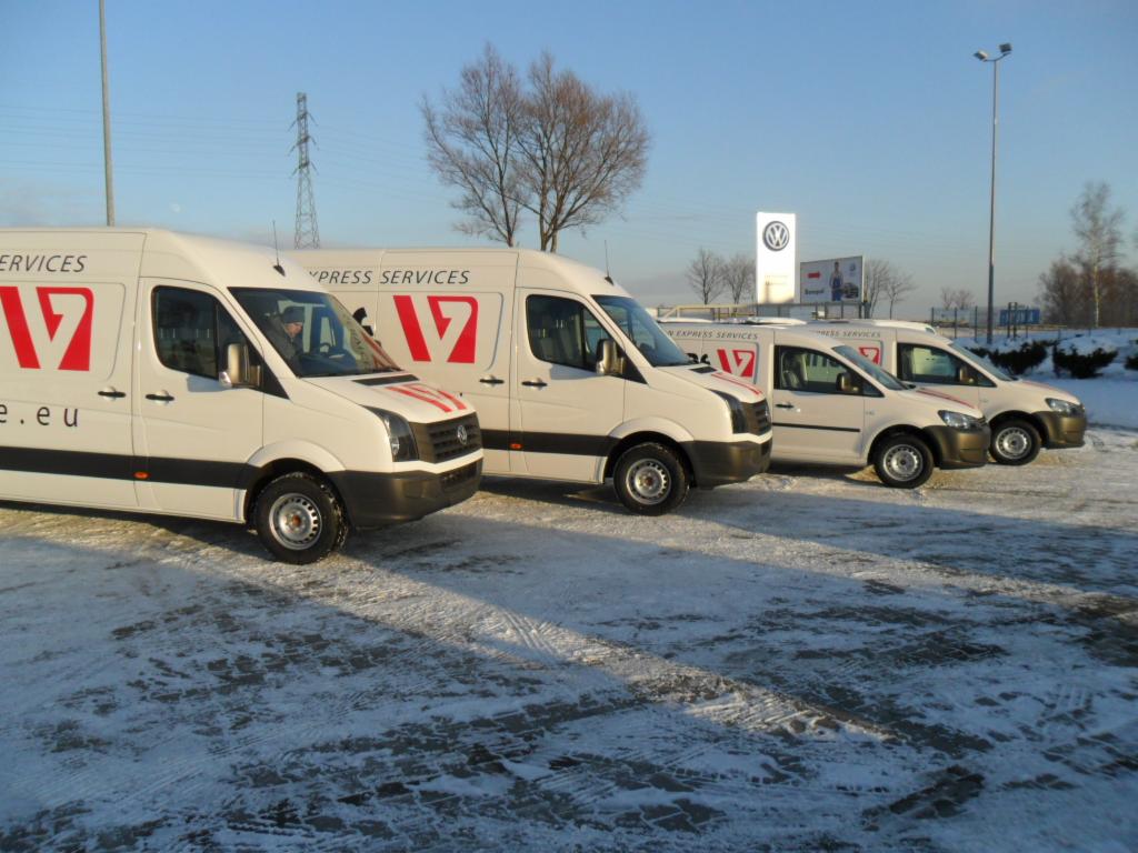 Flota białych samochodów dostawczych z logo firmy zaparkowana na ośnieżonym placu w słoneczny dzień.