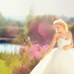 Panna młoda w białej sukni z gorsetem, pozuje na tle malowniczego krajobrazu z jeziorem i kwitnącymi kwiatami, oświetlona ciepłym światłem słonecznym.