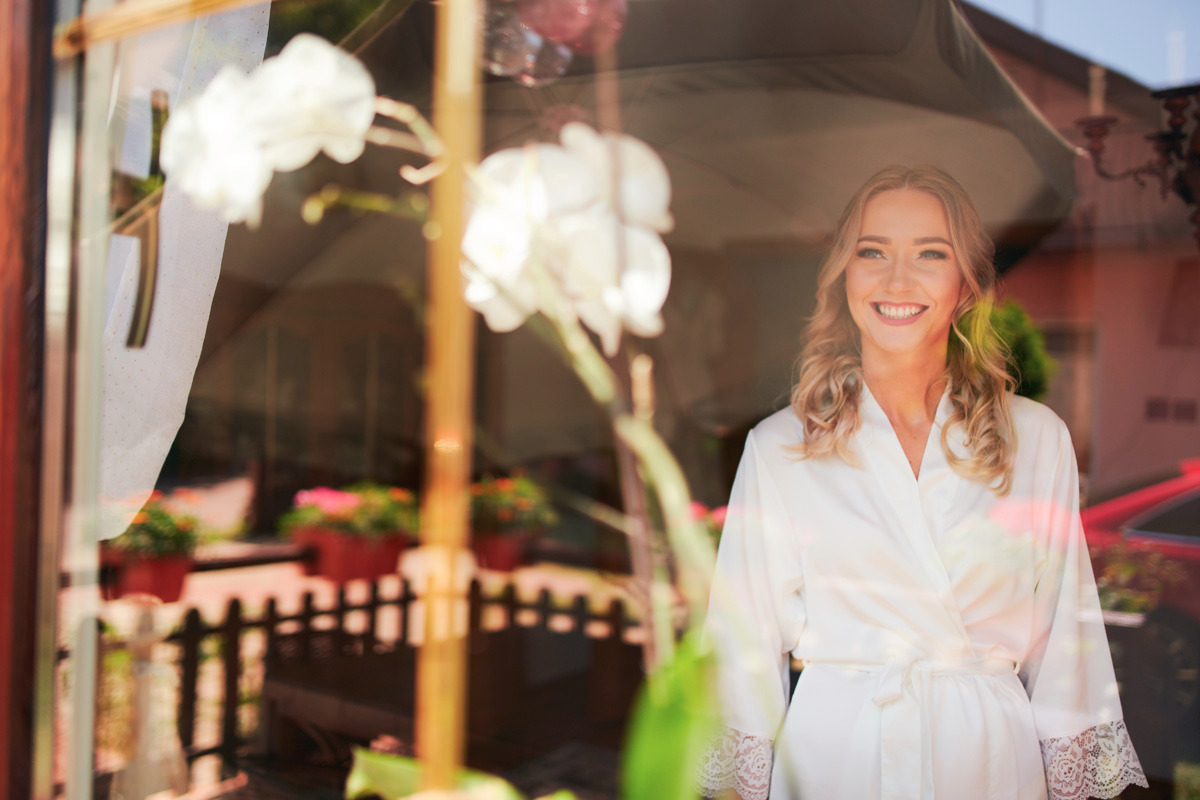 Uśmiechnięta panna młoda w białym szlafroku z koronkowymi wykończeniami, widziana przez okno z odbiciami kwiatów i otoczenia.