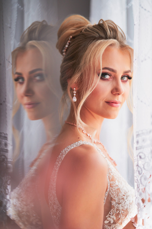 Portret panny młodej w sukni ślubnej z koronkowymi zdobieniami, z upiętymi blond włosami i delikatnym makijażem, z odbiciem w szybie.