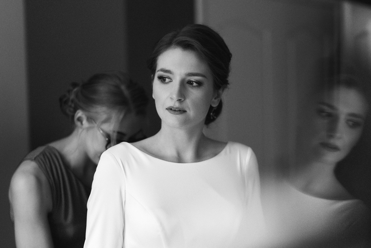 Czarno-białe zdjęcie panny młodej w prostej, białej sukience, spoglądającej w bok, z wizażystką poprawiającą jej fryzurę w tle i rozmytym odbiciem w lustrze.