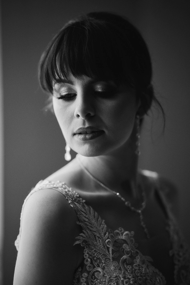 Portret panny młodej w czerni i bieli, z delikatnym makijażem, spuszczonym wzrokiem, w bogato zdobionej sukni z koronkowym haftem, naszyjnikiem i kolczykami z kryształkami.