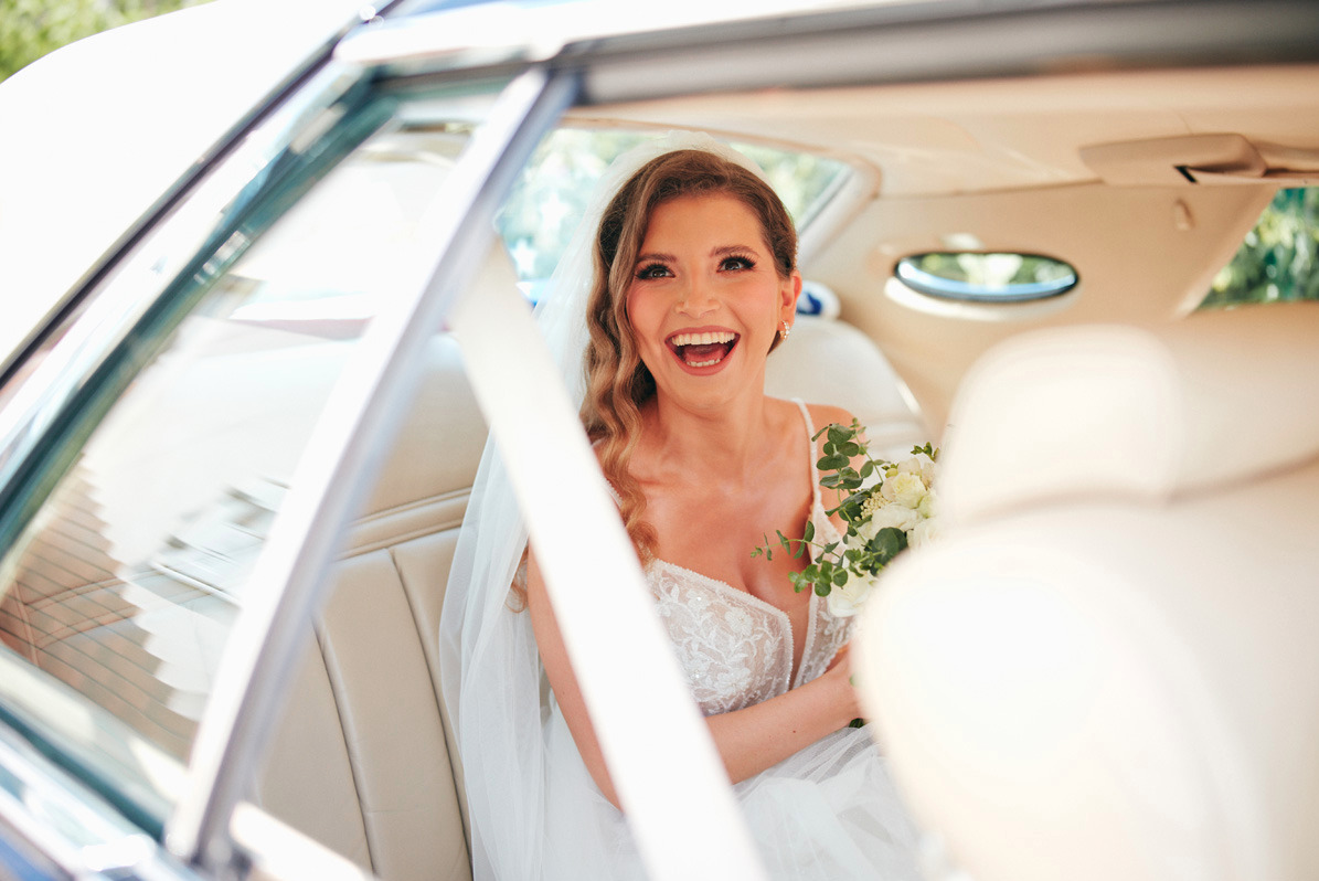 Uśmiechnięta panna młoda w sukni ślubnej z bukietem, siedząca w limuzynie, widok przez otwarte drzwi.