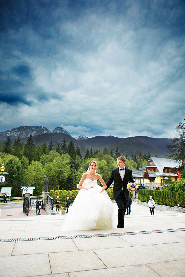 Para młoda schodzi po schodach na tle górskiego krajobrazu i zachmurzonego nieba, panna młoda w sukni bez ramiączek, pan młody w czarnym smokingu z muchą, trzyma bukiet fioletowych kwiatów.