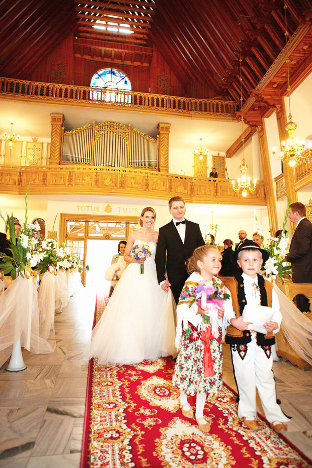 Para młoda w kościele, prowadzona przez dzieci ubrane w stroje ludowe, na tle drewnianego wnętrza z organami i czerwonym dywanem.