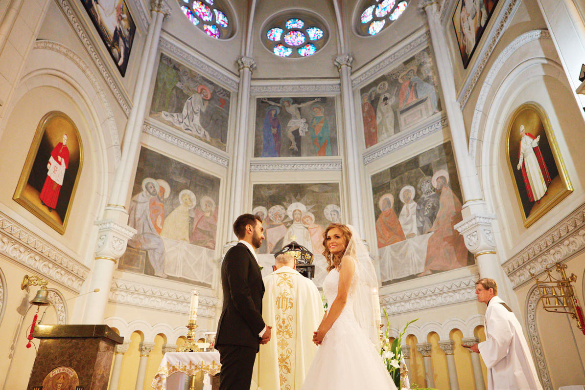 Para młoda podczas ceremonii ślubnej w bogato zdobionym kościele z freskami i witrażami, ksiądz w ornacie, świece na ołtarzu.