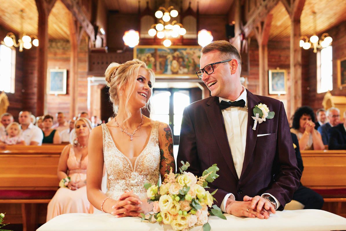 Uśmiechnięta para młoda w kościele, panna młoda w sukni z koronkowymi zdobieniami i tatuażem na ramieniu, pan młody w bordowym garniturze z muchą, bukiet na pierwszym planie, drewniane wnętrze...