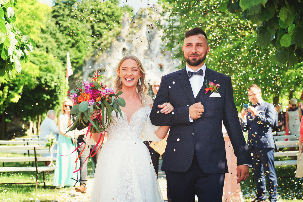 Uśmiechnięta para młoda, otoczona deszczem konfetti, idzie alejką w plenerze; Panna młoda trzyma bukiet z pomarańczowymi i różowymi kwiatami, a pan młody ma na sobie granatowy garnitur z muszką.