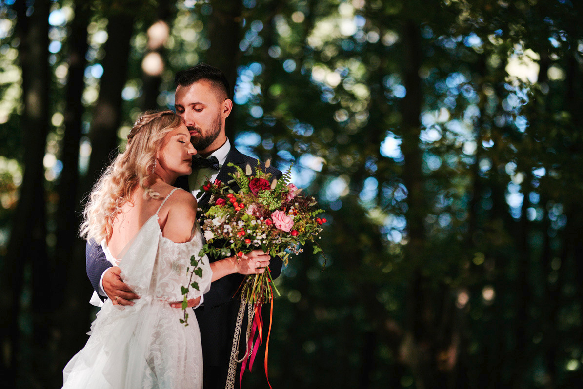 Para młoda w leśnej scenerii: panna młoda w koronkowej sukni z odkrytymi ramionami trzyma bukiet z różowych i czerwonych kwiatów, pan młody w ciemnym garniturze obejmuje ją czule.