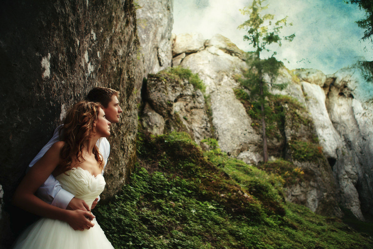Para młoda przytulona do skały, panna młoda w sukni bez ramiączek, pan młody w białej koszuli, w tle skalisty krajobraz z zielenią.