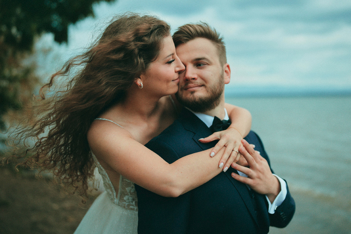 Para młoda w objęciach na tle jeziora; panna młoda z burzą falujących włosów w sukni z koronkowym dekoltem, całuje pana młodego w ciemnym garniturze z muchą.