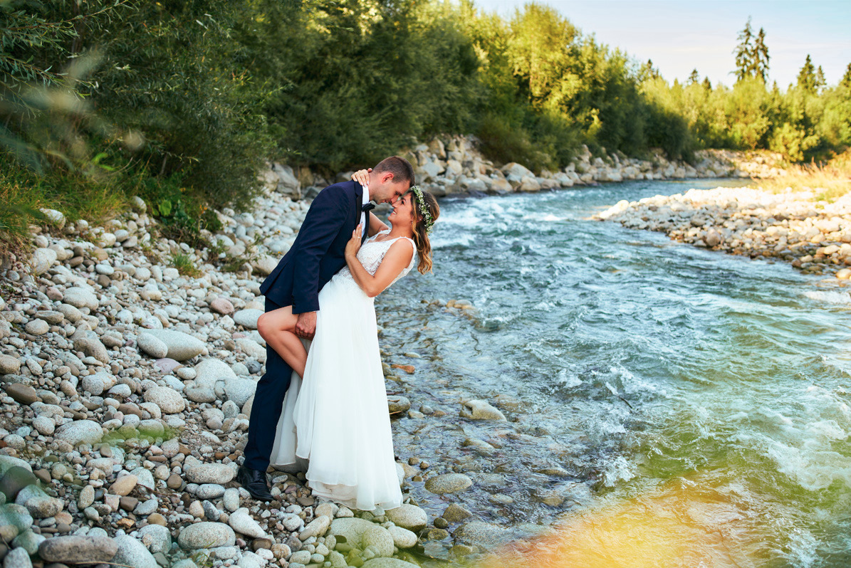 Para młoda w objęciach na tle górskiego potoku; panna młoda w białej sukni i wianku, pan młody w granatowym garniturze, oboje uśmiechnięci.