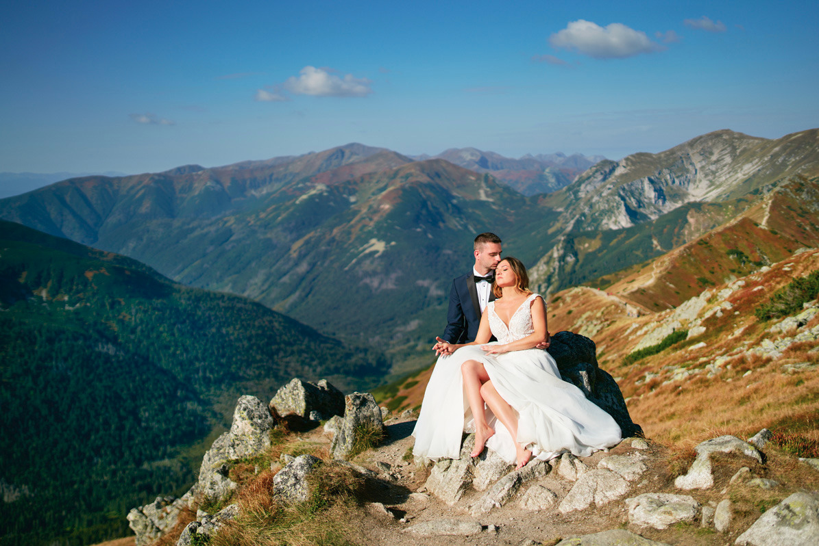 Para młoda w strojach ślubnych siedzi na skalistym szczycie góry z widokiem na rozległą panoramę górską w słoneczny dzień.