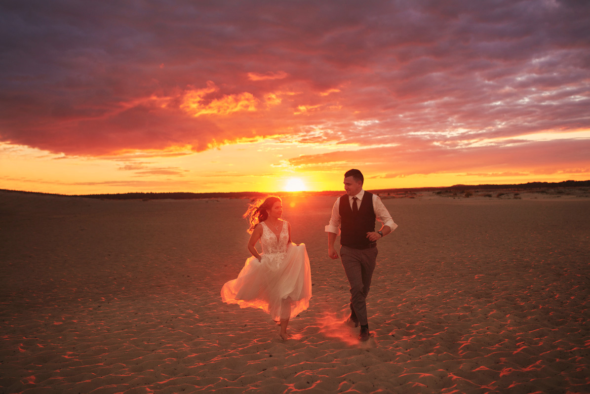 Para młoda biegnie po piaszczystej plaży w Myszkowie podczas zachodu słońca. Panna młoda w białej sukni z koronkową górą, pan młody w kamizelce i spodniach w kratę. Pomarańczowe niebo z chmurami...