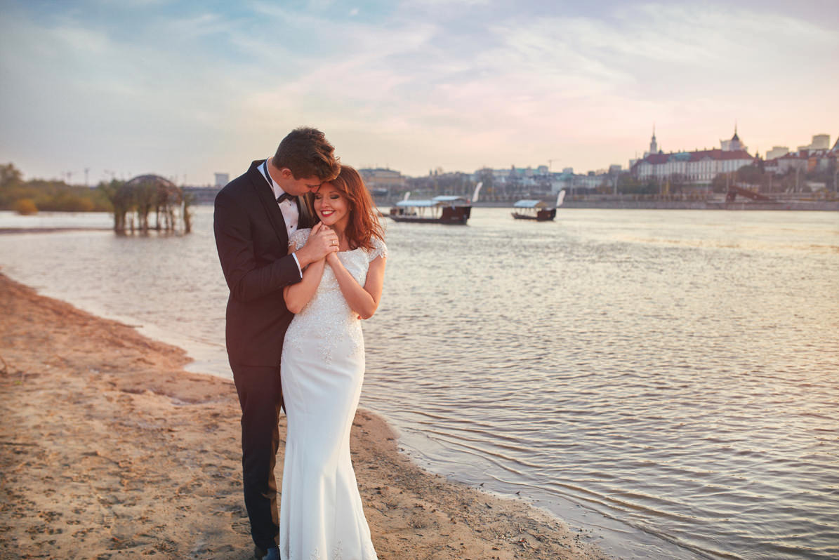 Para młoda na piaszczystej plaży nad rzeką z panoramą miasta w tle, on w czarnym smokingu, ona w białej sukni ślubnej z koronkowymi rękawami, trzymają się za ręce i uśmiechają.