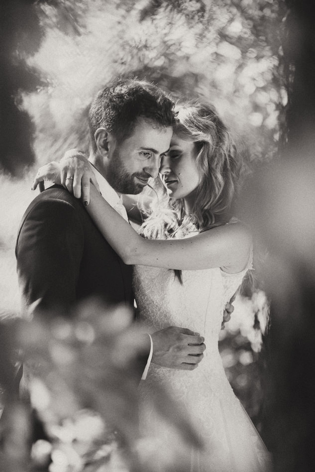 Czarno-białe zdjęcie pary młodej obejmującej się w plenerze, panna młoda w koronkowej sukni, pan młody w garniturze, delikatne rozmycie tła.