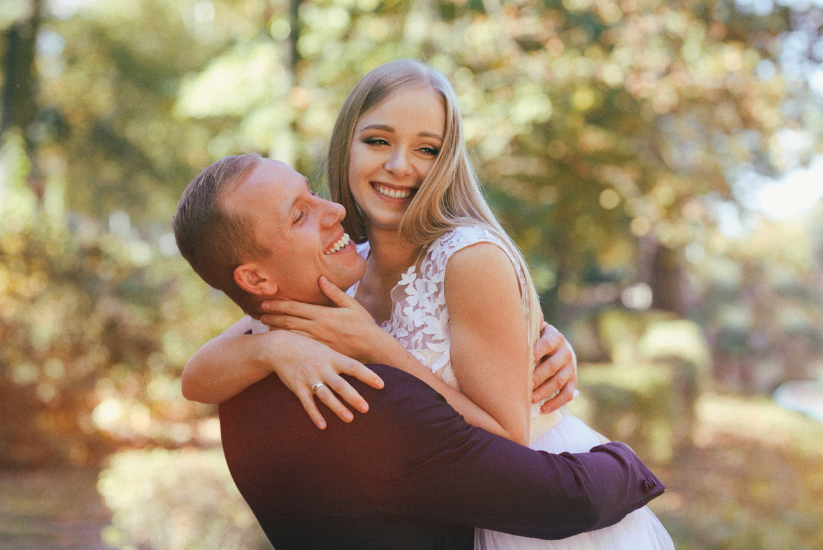 Uśmiechnięta para w objęciach na tle drzew, panna młoda w białej sukience z koronkową górą, widoczny pierścionek na palcu mężczyzny.