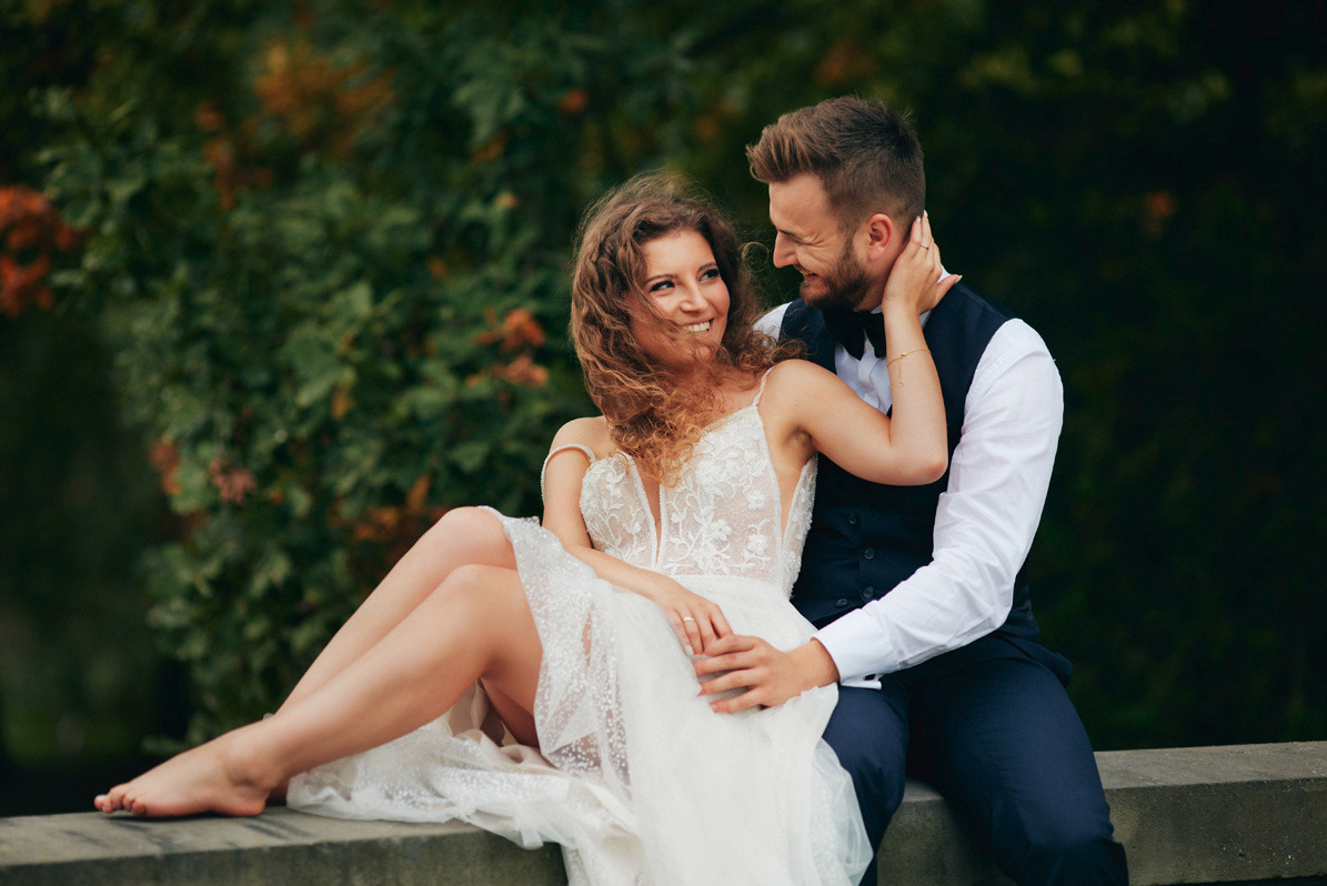 Uśmiechnięta panna młoda w koronkowej sukni siedzi na murku, obejmowana przez pana młodego w muszce, na tle zieleni.