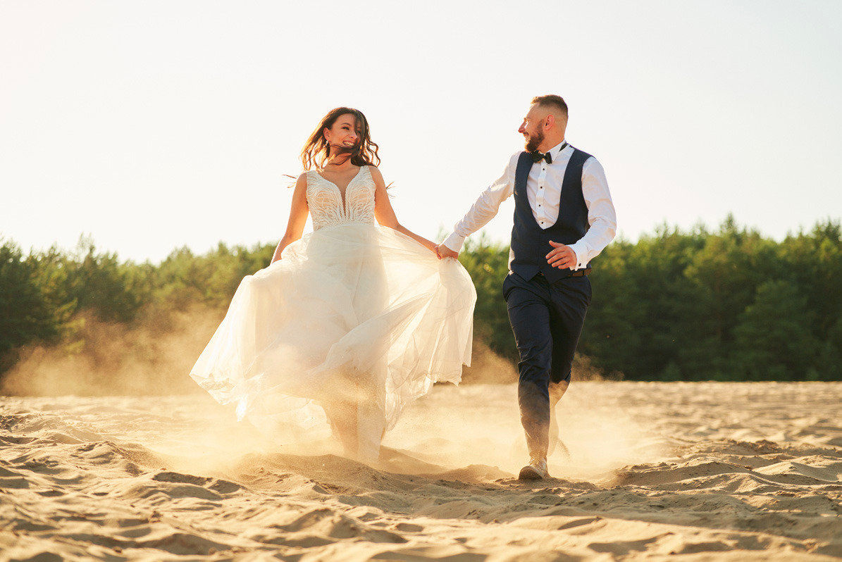 Para młoda biegnie po piaszczystej plaży, unosząc tumany piasku; panna młoda w zwiewnej sukni z koronkową górą, pan młody w granatowej kamizelce i muszce, w tle las iglasty.