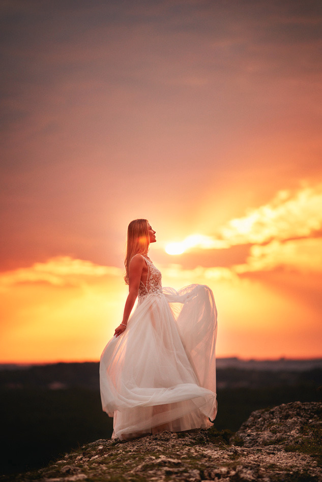 Panna młoda w białej sukni z koronkową górą, stojąca na skalistym wzgórzu na tle zachodzącego słońca, które tworzy pomarańczowo-różowe niebo.