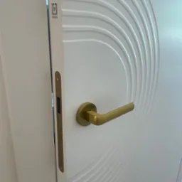 Białe skrzydło drzwiowe z dekoracyjnymi frezami w kształcie łuków i złotą klamką, widoczny zamek wpuszczany oraz logo producenta.