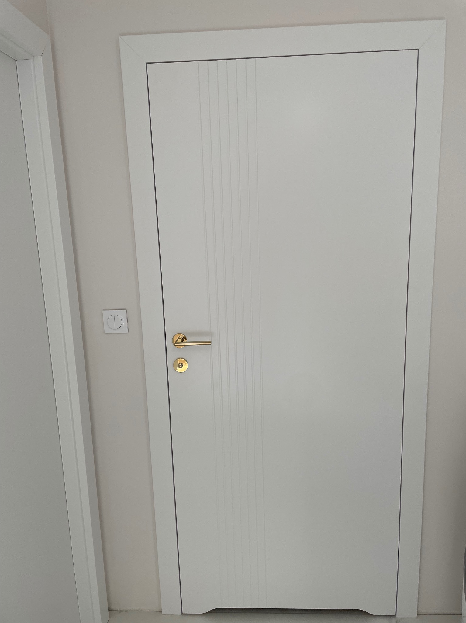 Białe skrzydło drzwiowe z delikatnymi pionowymi frezami, zamontowane w białej ościeżnicy, z złotą klamką i zamkiem, widoczne na tle jasnej ściany i fragmentu sąsiednich drzwi.