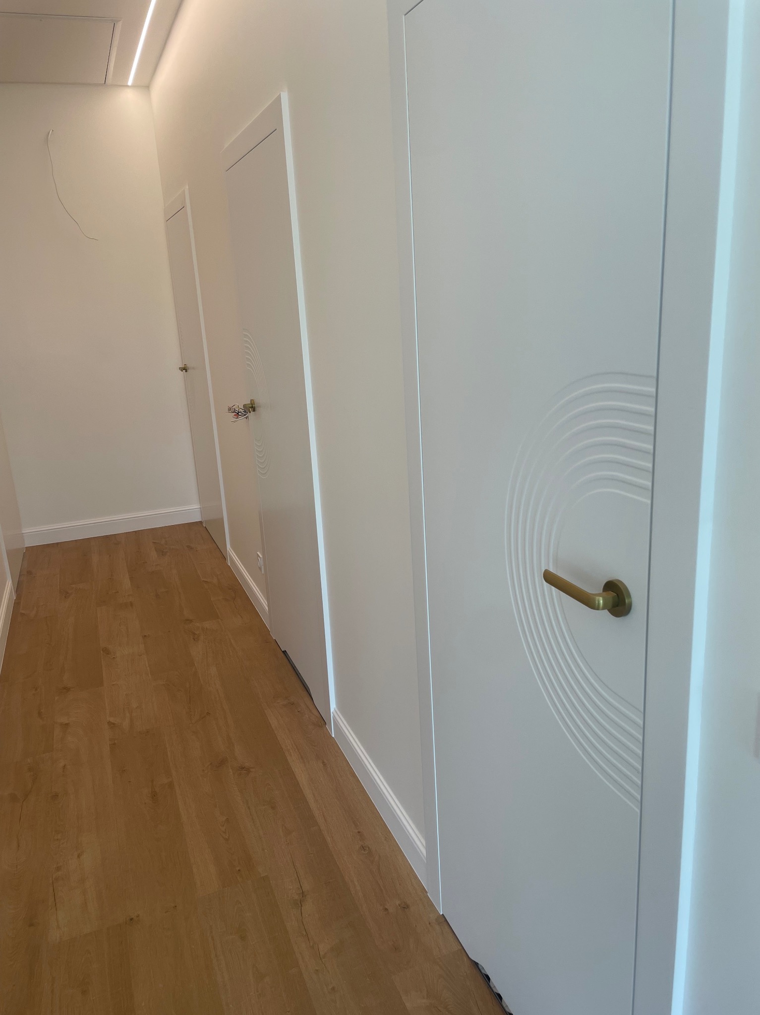 Korytarz z trzema białymi skrzydłami z dekoracyjnym motywem w kształcie łuku i złotymi klamkami, widoczne na drewnianej podłodze.