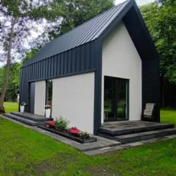 Nowoczesny, parterowy dom z dwuspadowym dachem z blachy na tle zielonego ogrodu, z dekoracyjnymi donicami przy wejściu i schodami z kamiennych płyt.