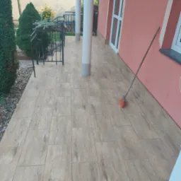 Ułożona podłoga z paneli imitujących drewno na zewnątrz budynku, z widoczną szczotką opartą o różową ścianę.