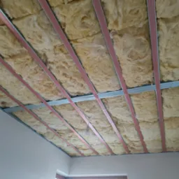 Rama z profili metalowych podwieszanego sufitu wypełniona izolacją z wełny mineralnej, widoczne fragmenty ścian i żarówka na kablu.