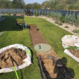 Przygotowanie podłoża pod trawnik z rolki w Białystoku: wykopany pas ziemi, widoczne worki z ziemią, narzędzia ogrodnicze oraz taczka z trawą w rolkach.