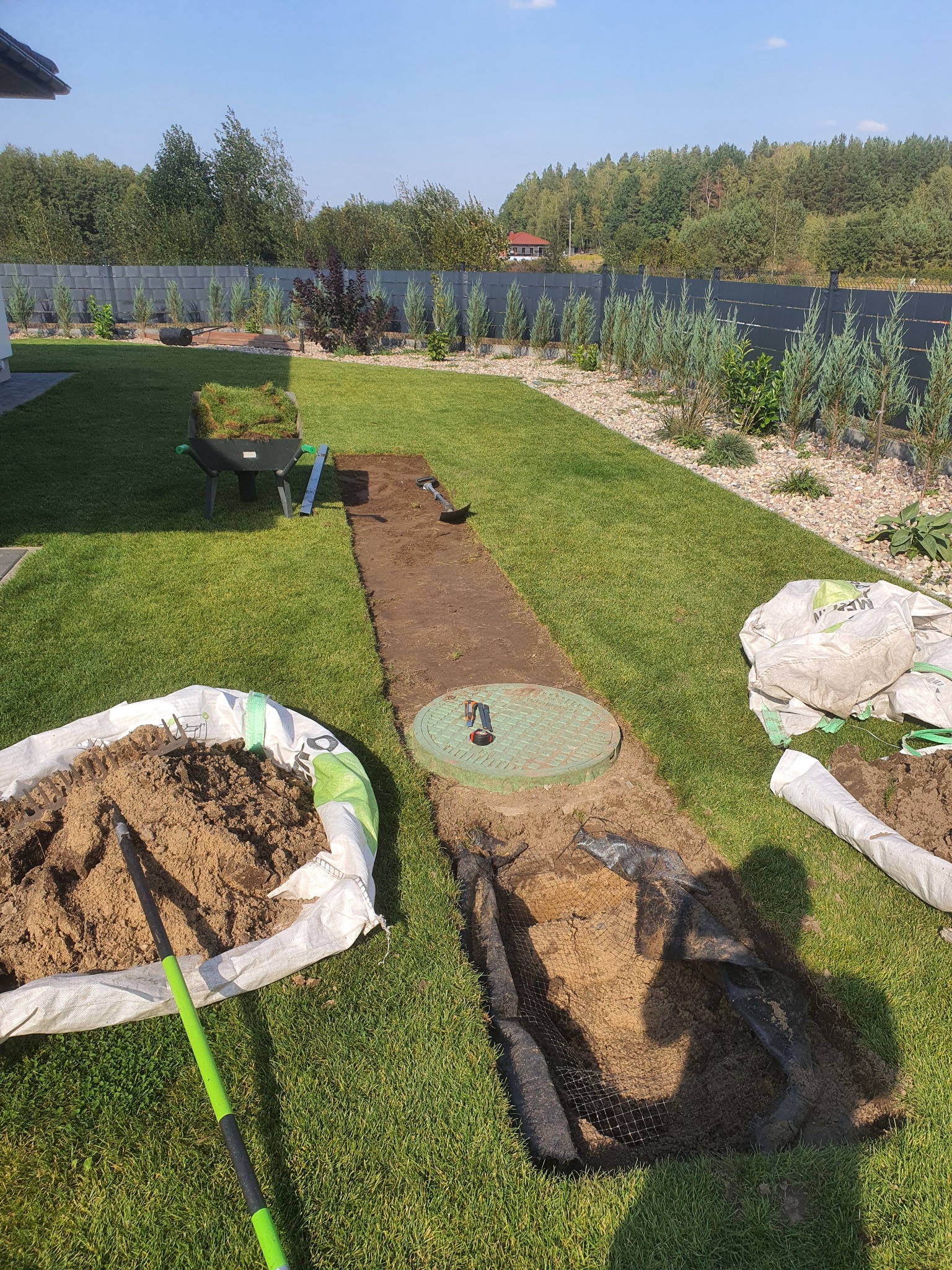 Przygotowanie podłoża pod trawnik z rolki w Białystoku: wykopany pas ziemi, widoczne worki z ziemią, narzędzia ogrodnicze oraz taczka z trawą w rolkach.