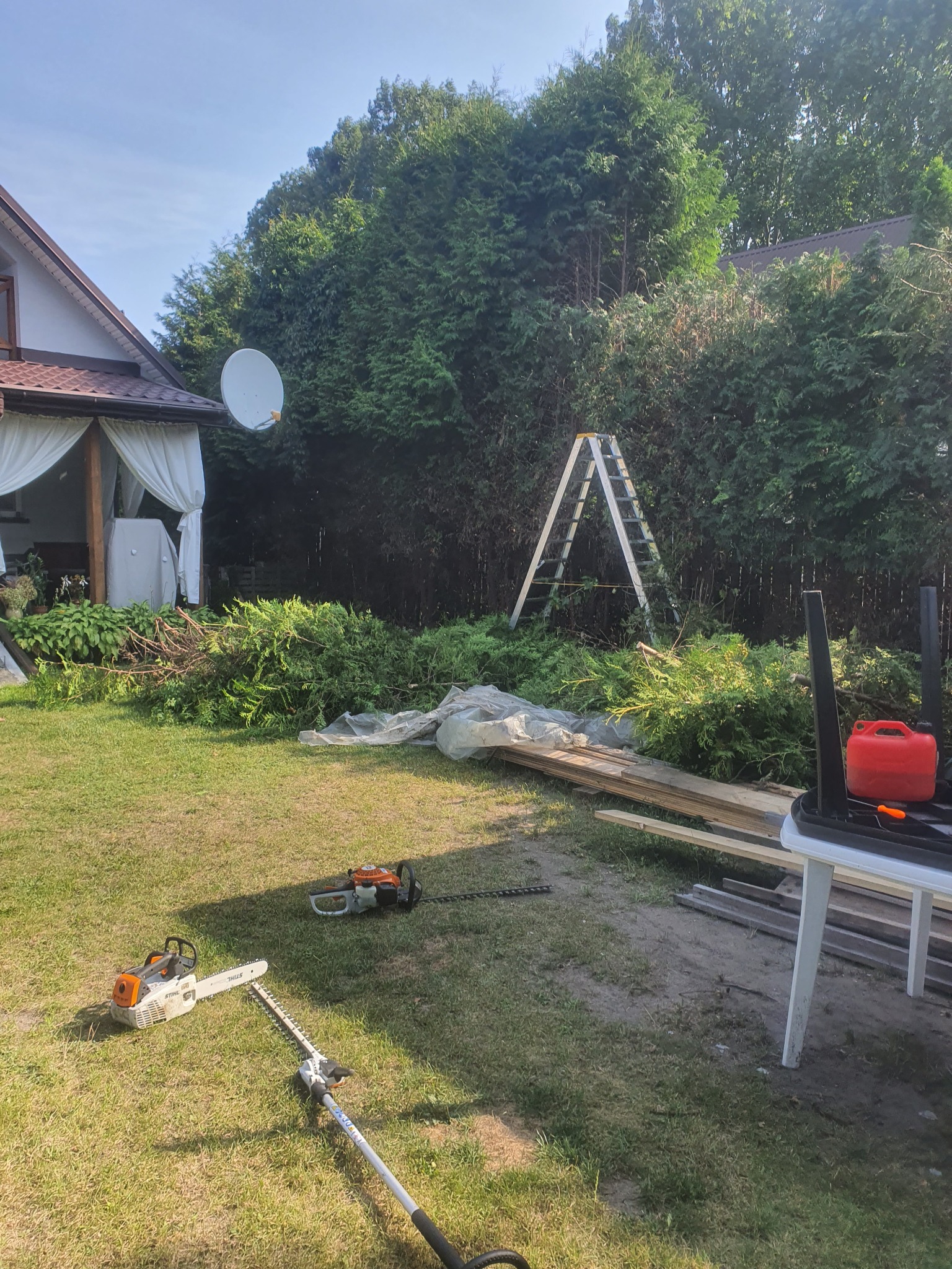 Po pracy: Przycięte gałęzie iglaków leżą na trawniku obok drabiny, pilarki spalinowej i nożyc do żywopłotu; w tle dom z białą altaną i zielone krzewy.