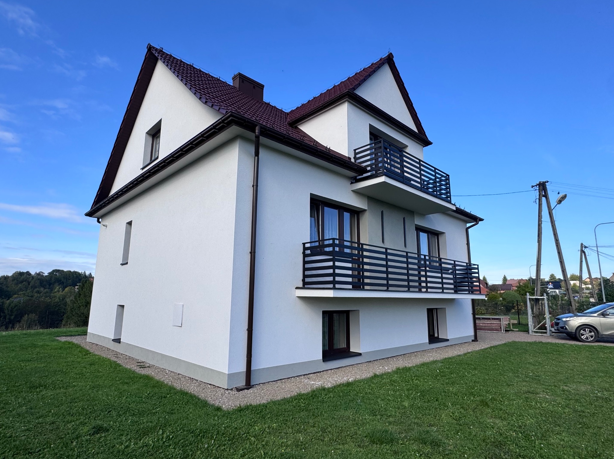 Dwupiętrowy dom z białymi ścianami i ciemną dachówką, balkony z czarną balustradą, otoczony zielonym trawnikiem i błękitnym niebem. Widok z perspektywy trawnika.