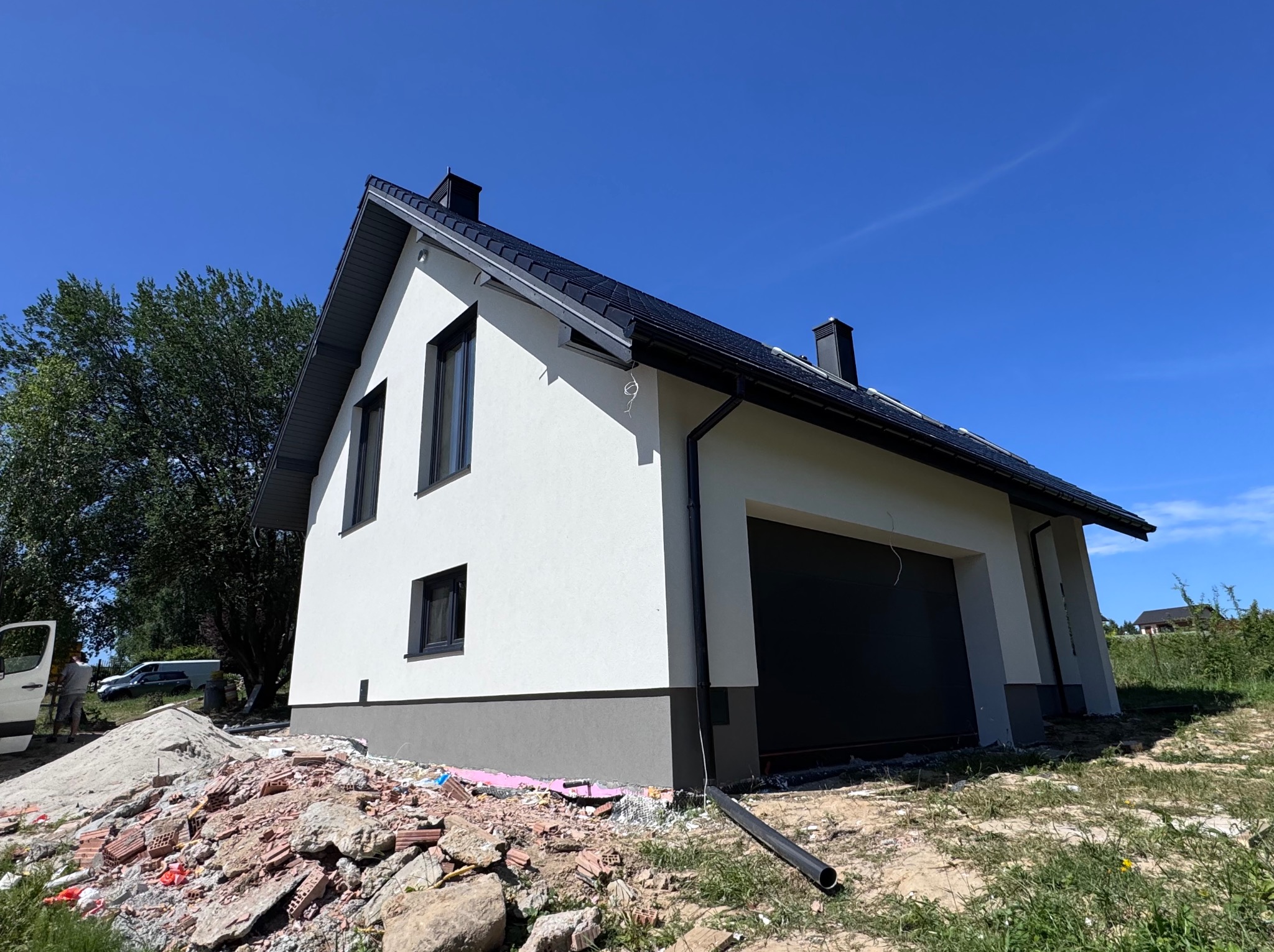 Biały dom z szarą podmurówką i czarnym dachem w słoneczny dzień. Widoczne gruz i materiały budowlane, sugerujące prace wykończeniowe elewacji.