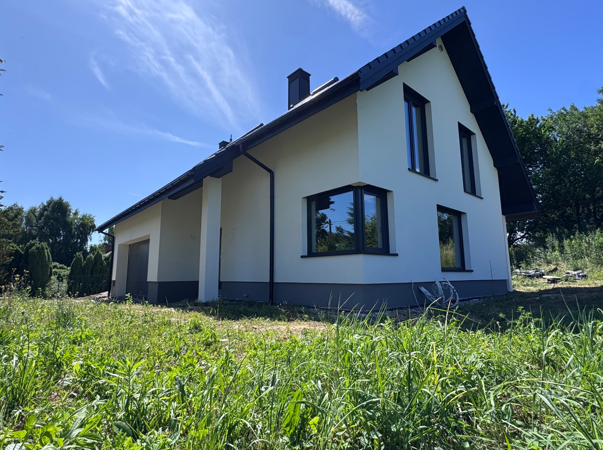 Nowoczesny dom jednorodzinny z jasną elewacją i ciemną stolarką okienną, widoczny z poziomu trawnika w słoneczny dzień. Minimalistyczny design, dwuspadowy dach.