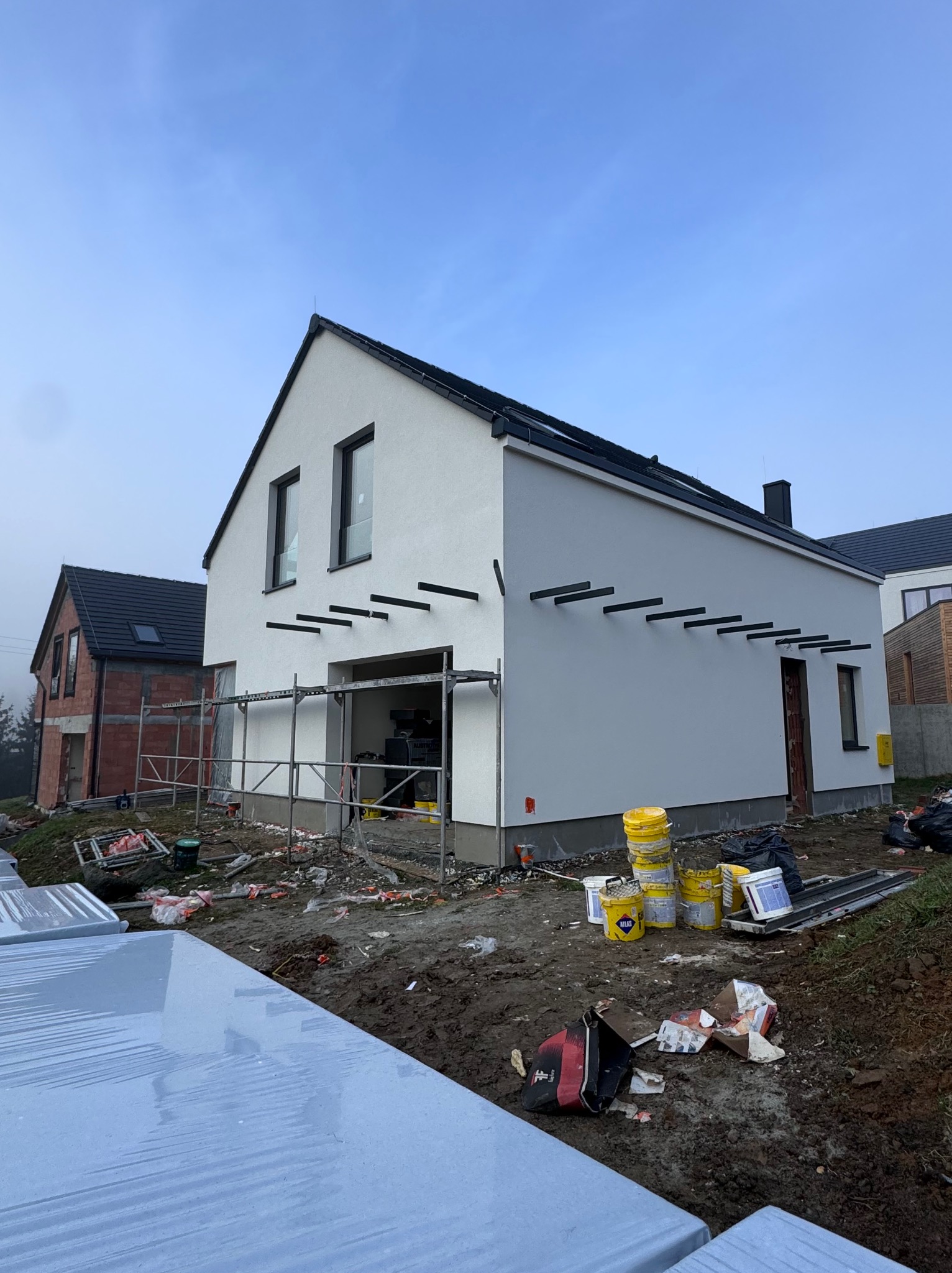 Biały dom jednorodzinny z czarnymi elementami dekoracyjnymi, w trakcie prac wykończeniowych elewacji. Widoczne rusztowania, farby i narzędzia budowlane.