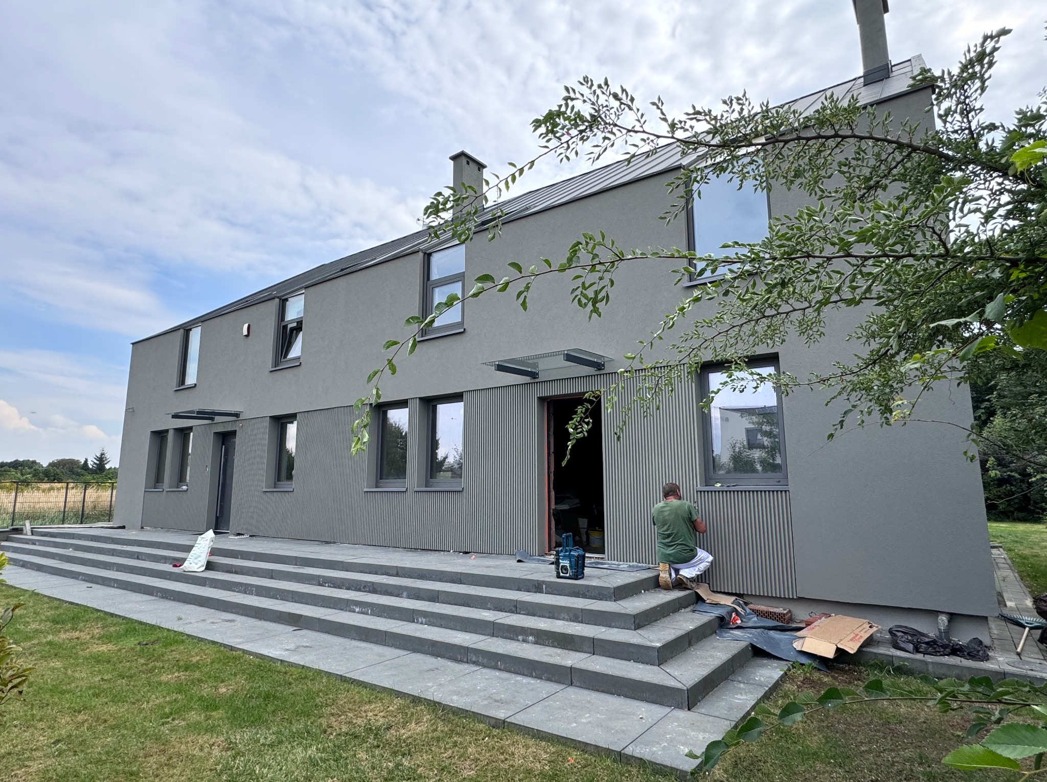Szary, dwukondygnacyjny dom z płaskim dachem i schodami wejściowymi. Mężczyzna w zielonej koszulce maluje elewację przy drzwiach wejściowych. Widoczne narzędzia pracy.