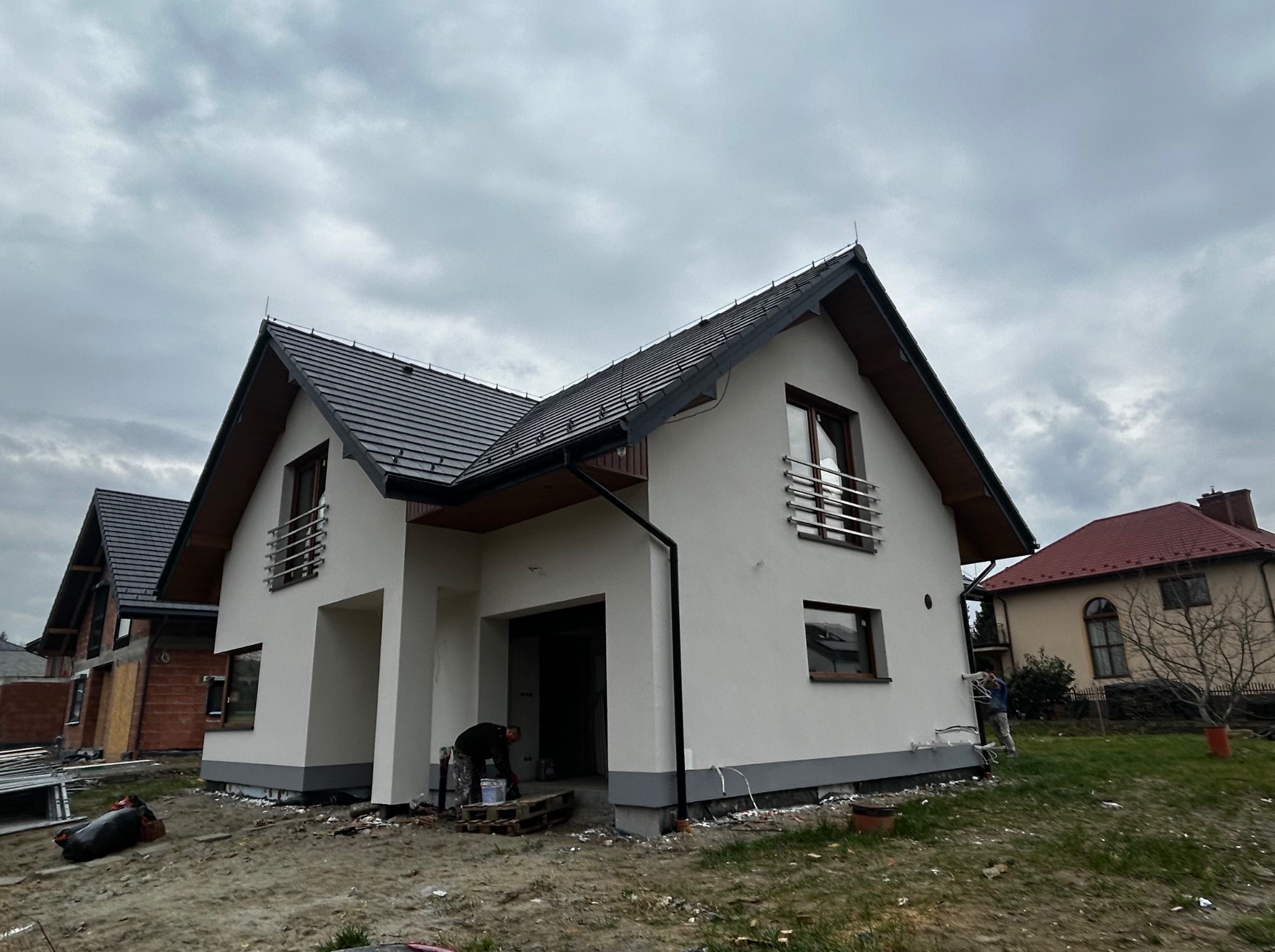 Nowoczesny dom jednorodzinny z białą elewacją w trakcie prac wykończeniowych. Widoczny pracownik przy wejściu do garażu i drugi malujący ścianę. Pochmurne niebo.