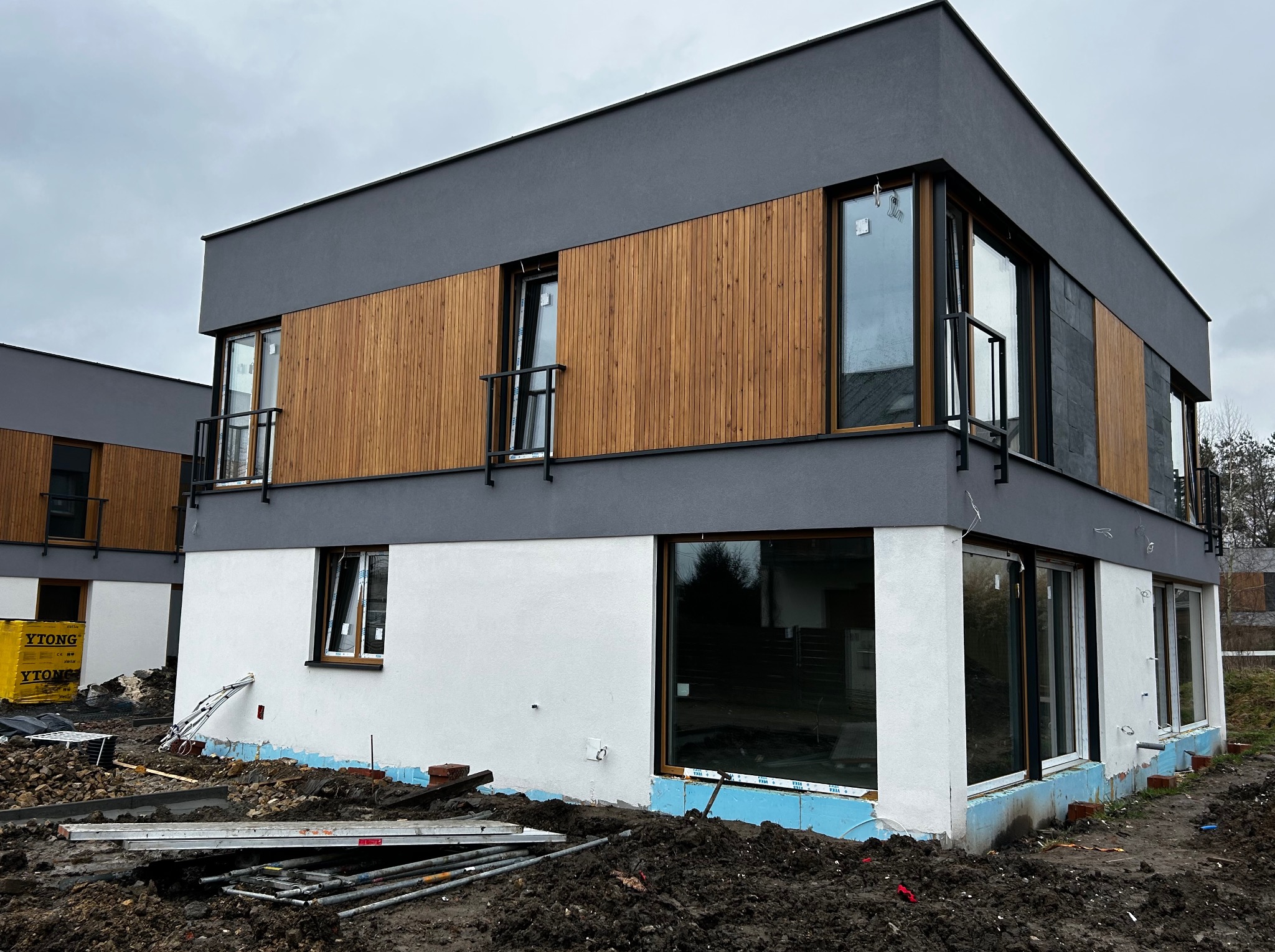 Nowoczesny dom w trakcie wykańczania elewacji: biały tynk, drewniane panele, grafitowe elementy. Widoczne materiały budowlane i teren wokół budynku.
