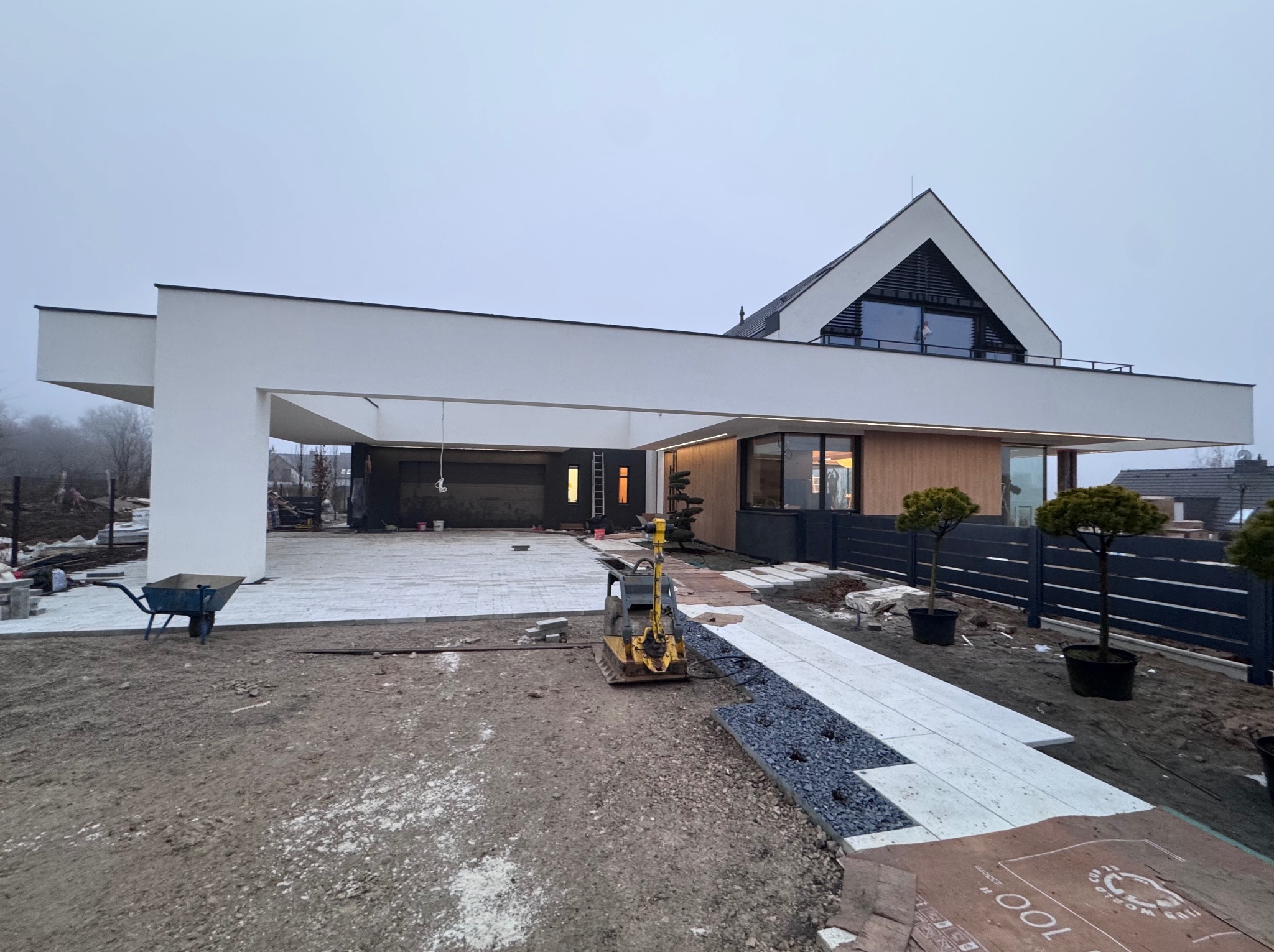 Nowoczesny dom jednorodzinny z elementami drewnianymi i białymi, płaskim dachem nad garażem. W tle niebo, na pierwszym planie prace brukarskie i zagęszczarka.